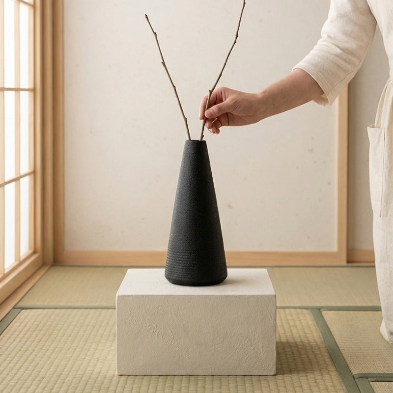 Hand arranging dried branches in japanese vintage vase black displayed on white pedestal over traditional tatami