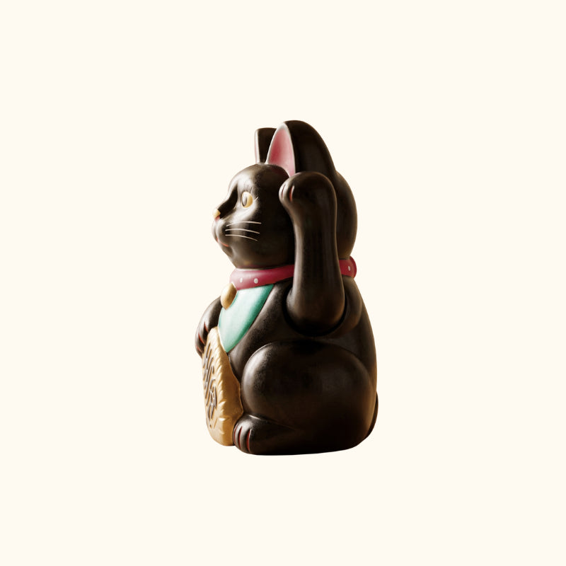 Side profile of a black lucky Maneki Neko cat statue with a raised paw, green bib and gold coin.