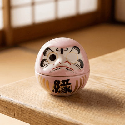 Pastel pink daruma doll with determined expression resting on wooden surface in minimalist Japanese setting with soft natural light