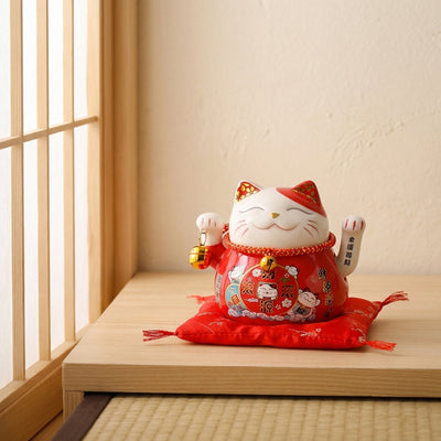 Red maneki neko statue with fringed satin cushion and gold bell charm, positioned on a low wooden step beside a shoji screen door on a tatami floor