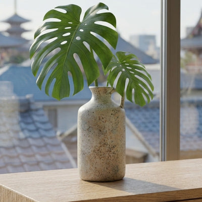Textured vintage ikebana vase with speckled glaze holding fresh monstera leaves on shelf overlooking japanese temple rooftops