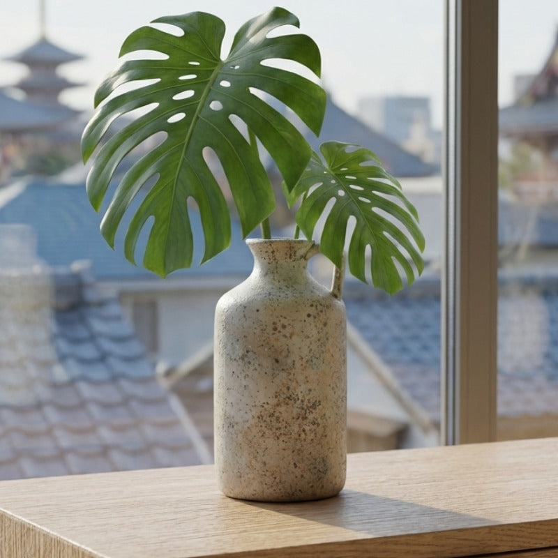 Textured vintage ikebana vase with speckled glaze holding fresh monstera leaves on shelf overlooking japanese temple rooftops