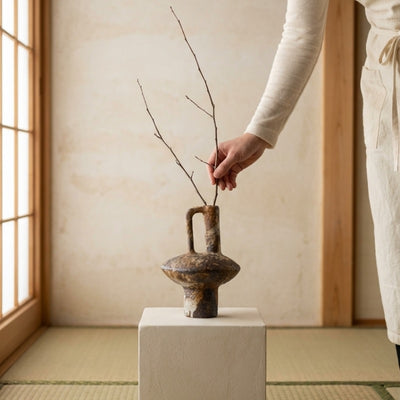 Hand arranging minimalist branch in authentic wabi sabi vase long displayed on white pedestal over tatami