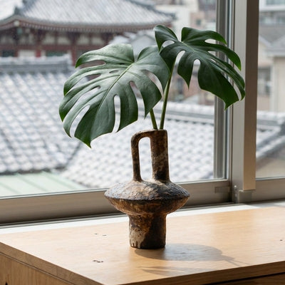Sculptural wabi sabi vase holding fresh monstera leaf by window overlooking traditional japanese temple rooftops